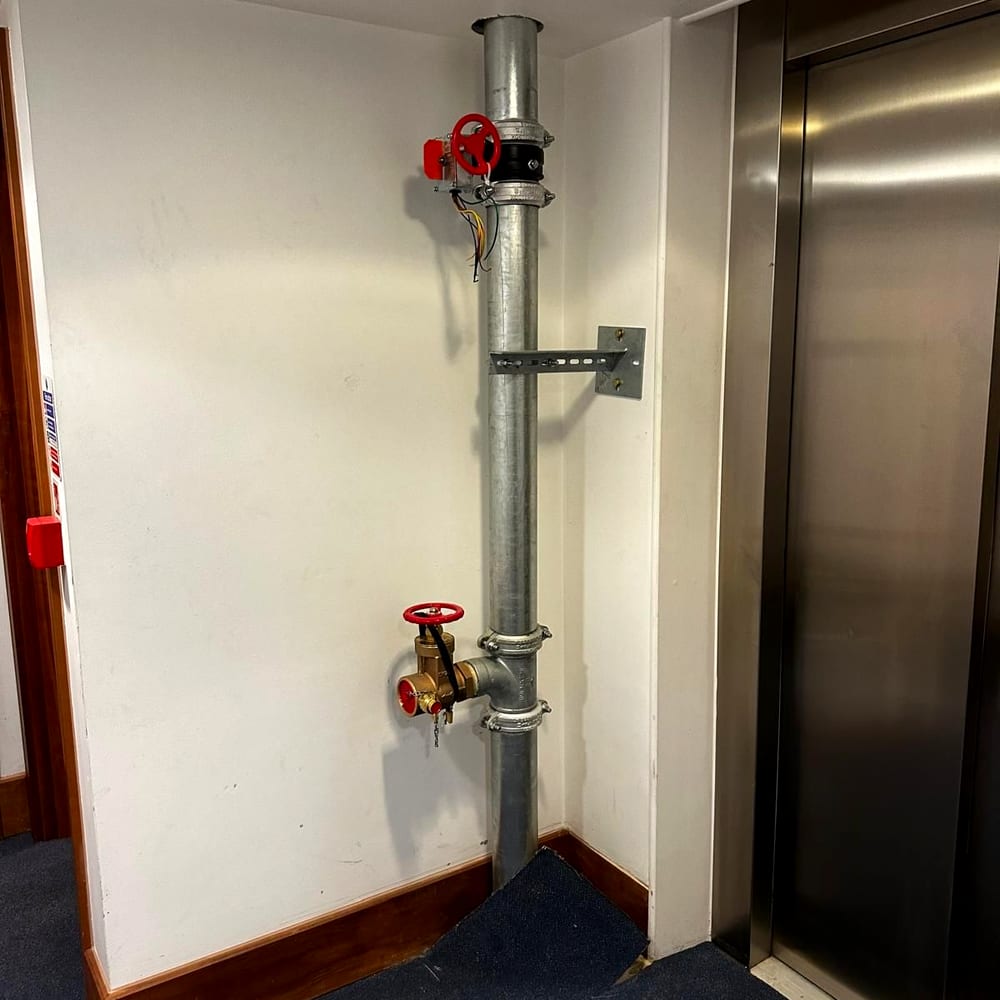 A dry riser landing valve with its red handwheel on a galvanised riser beside a lift in a residential lobby